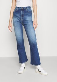 Jeans acampanados de mezclilla azul de cintura alta con un acabado deslavado. Incluyen cierre frontal de botones y cinco bolsillos. Combinados con zapatillas blancas.