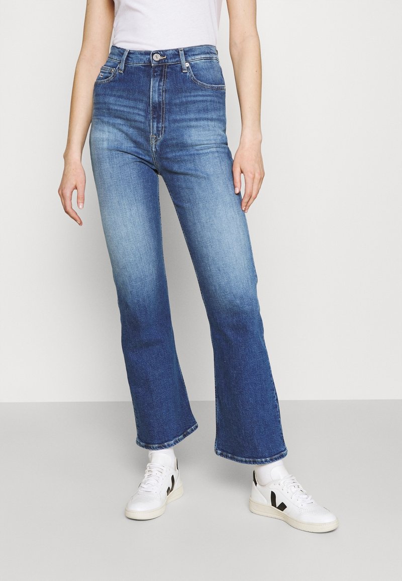 Jeans acampanados de mezclilla azul de cintura alta con un acabado deslavado. Incluyen cierre frontal de botones y cinco bolsillos. Combinados con zapatillas blancas.