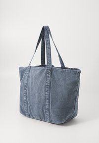 Stor stor blåvasket denim tote bag med to lange håndtak, stående oppreist på en lys bakgrunn.