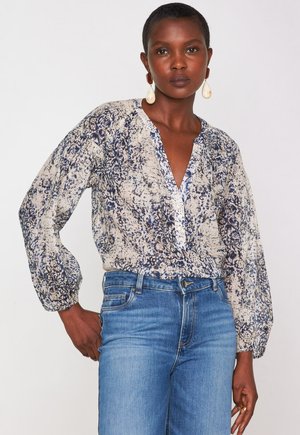 Leichte, cremefarbene Bluse mit blauem Blumenmuster, tiefem V-Ausschnitt und gerafften Ärmeln. Kombiniert mit blauen Jeans und schlichten Ohrringen.