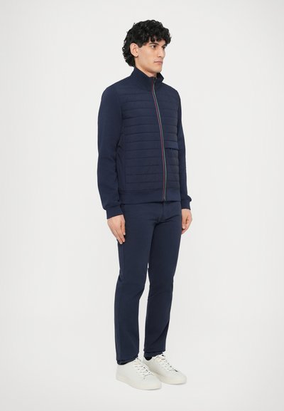 Tumesinine lukuga jope, millel on tepitud esipaneelid, kõrge krae ja mitmevärviline lukk. Sobitatud vastavate pükste ja valgete tossudega.