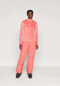 Różowy polarowy top na zamek i pasujące różowe spodnie narciarskie, wykonane z miękkiego, ocieplanego materiału. Całość dopełniają czarne buty narciarskie.