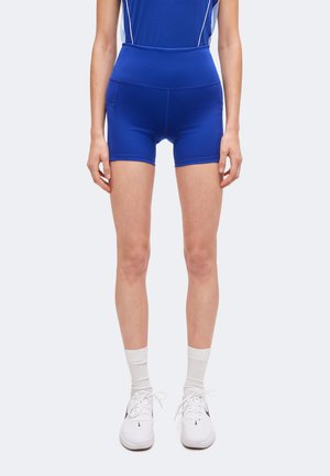 Persoon met een mouwloos blauw sporttop, bijpassende blauwe high-waisted sportshort, witte sokken en witte sportschoenen, rechtop staand.