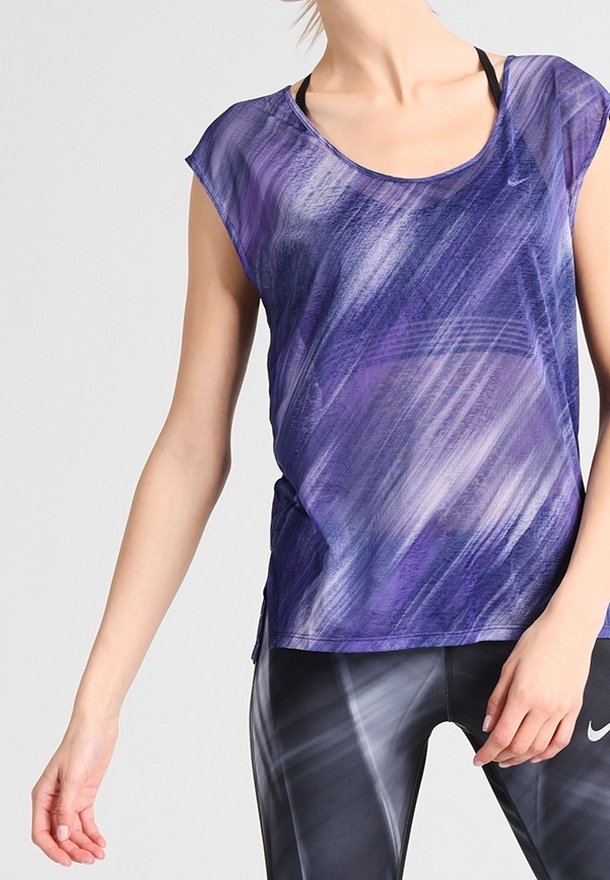 Vrouw die een paarse, patroonloze, mouwloze sporttop draagt en zwarte Nike sportleggings met witte strepen, staand met één arm ontspannen.