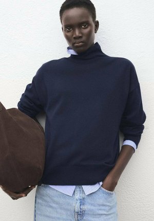 Pull en tricot marine avec un col montant, superposé à une chemise rayée bleu clair et blanche, associé à un jean en denim clair et un sac marron.