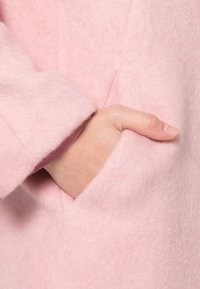 Abrigo de lana rosa con una textura suave y esponjosa, que presenta una mano en un bolsillo lateral y un forro en contraste visible en el borde de la manga.