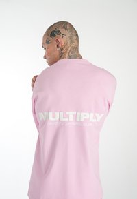 Růžové bavlněné tričko s velkým bílým logem "MULTIPLY" a menším textem na zádech. Textilie vypadá hladce a lehce.