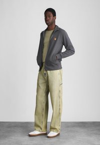 Gray zip-up jacket with a small orange emblem, layered over a green shirt. Light green striped wide-leg jeans and white sneakers.