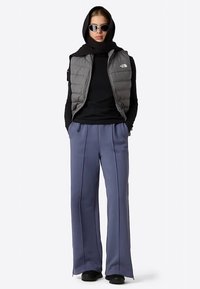 Kamizelka puchowa w kolorze szarym na czarnej koszuli z długim rękawem, zestawiona z szerokimi niebieskimi spodniami. Dopełniona czarną chustą na głowę i okularami przeciwsłonecznymi.