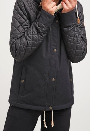 Veste matelassée sombre avec boutons-pression et cordon de serrage, portée par une personne ajustant le col, montrant le haut du corps et les mains.