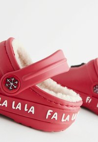 Röda gummiklumpor med en fluffig inredning, justerbar rem och vitt tryckt text som säger "FA LA LA LA." Snöflingeemblem på remmen.