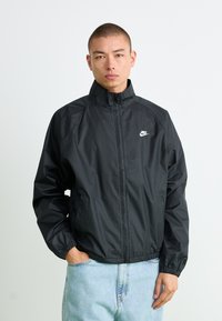 Mand iført en sort Nike zip-up jakke og lyseblå jeans, står med den ene hånd i lommen foran en ensfarvet hvid baggrund.