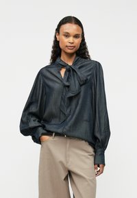 Tumša denima blūze ar sasienamo detaļu ap kaklu, garām piedurknēm un savilktiem aproču galiem, apvienota ar gaiši bežām biksēm. Gluda tekstūra un brīva piegriezuma.