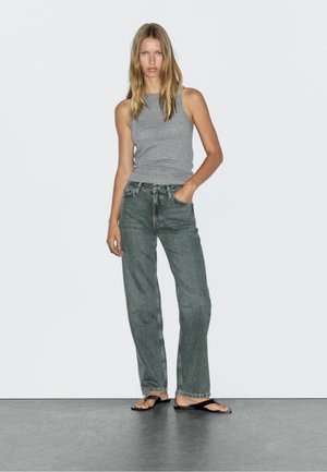 Grå linne, löst sittande mörkgröna jeans och svarta flip-flops. Modellen står på en slät, mörk yta med en neutral bakgrund.