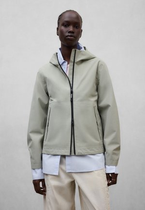 Persona con chaqueta con capucha de color gris claro con cremallera negra, camiseta blanca debajo y pantalones de color claro contra un fondo gris liso.