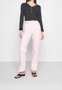 Schwarz gemusterte Bluse mit Knopfdetails am Ausschnitt, kombiniert mit hoch taillierten, hellrosa Hosen und schwarzen flachen Sandalen.
