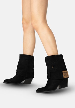 Benen die zwarte suède laarzen tot halverwege de kuit dragen met gestapelde hakken en een bruin patchdetail aan de buitenkant tegen een witte achtergrond.