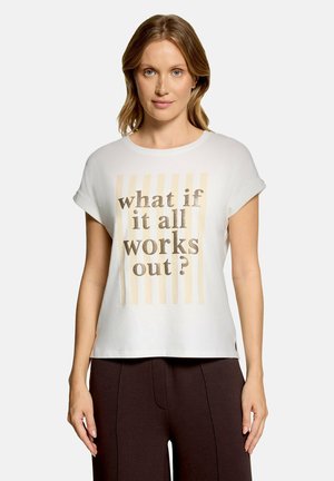 Kvinde med lysebrunt hår bærer hvid kortærmet skjorte med beige striber og teksten "what if it all works out?" kombineret med mørke bukser.
