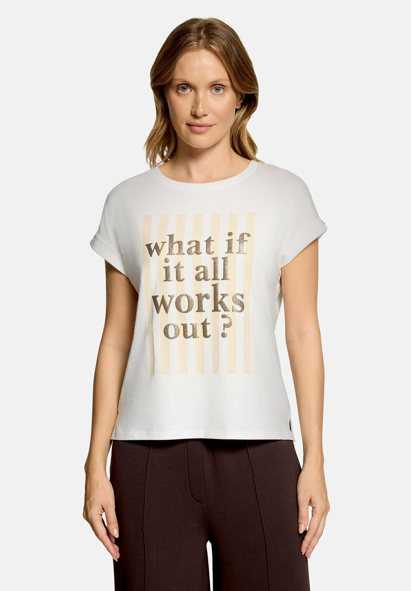 Femme aux cheveux châtain clair portant un t-shirt blanc à manches courtes avec des rayures beiges et le texte "what if it all works out?", associé à un pantalon foncé.