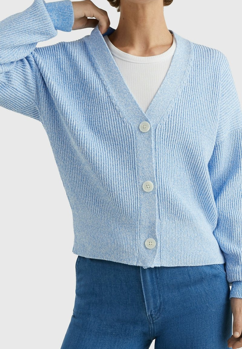 Personne portant un cardigan côtelé bleu clair à trois boutons, un haut blanc en dessous et un jean bleu, ajustant le col du cardigan d'une main.