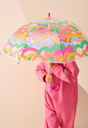 Enfant en combinaison de pluie rose tenant un parapluie transparent avec des rayures ondulées colorées, debout sur un fond beige clair.