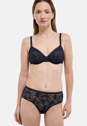 Femme portant un ensemble assorti de soutien-gorge en dentelle noire et de culottes taille haute, debout devant un fond blanc uni.
