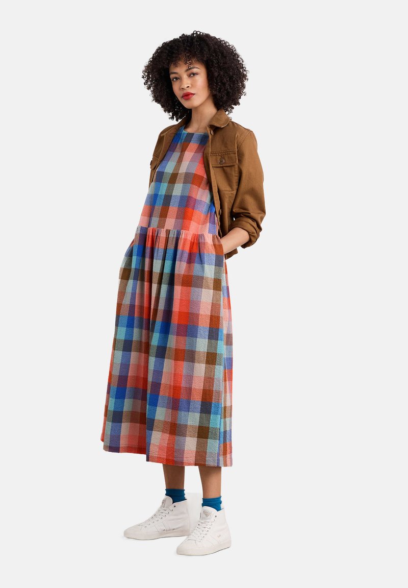 Maxi dress a scacchi colorati in tonalità di rosso, blu e marrone; presenta un colletto a barca, tasche e una gonna a strati. Abbinato a un giubbotto marrone e sneakers bianche.