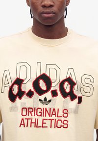 Νεαρό άτομο που φοράει κρεμ t-shirt Adidas Originals Athletics με μαύρα και κόκκινα κεντημένα γράμματα στην μπροστινή πλευρά.