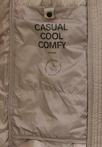 Etiqueta de cuidado de tejido beige que presenta "CASUAL COOL COMFY" y "S.OLIVER" en texto negro en negrita, con un gráfico de instrucciones de cuidado debajo.