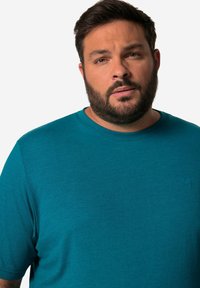 Tyrkysové tričko s kulatým výstřihem vyrobené z měkkého, texturovaného materiálu. Má krátké rukávy a nenápadný logo detail na hrudi.