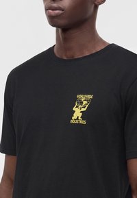 Νέος άνδρας που φοράει μαύρο T-shirt με κίτρινο γραφικό ατόμου που κρατάει μια υδρόγειο και το κείμενο "Worldwide Industries" στο στήθος.