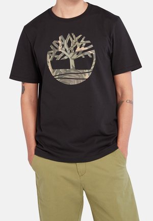 Sort bomulds-T-shirt med en rund camouflagemotiv af et træ i midten. Korte ærmer og en afslappet pasform. Båret med olivengrønne bukser.