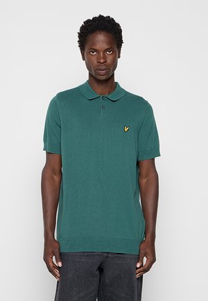 Mand iført en teal kortærmet polo med et lille gult fuglelogo på brystet, kombineret med mørke jeans, stående mod en ensfarvet baggrund.