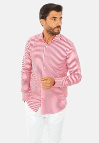 Camicia a righe rosse e bianche con maniche lunghe, colletto, e tessuto liscio; abbinata a pantaloni bianchi.