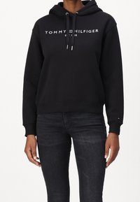 Schwarzer Kapuzenpullover mit weißem "TOMMY HILFIGER"-Text und Logo. Gerippte Bündchen und Saum. Getragen mit dunklen Jeans, die ein kleines Logo-Detail am Ärmel haben.