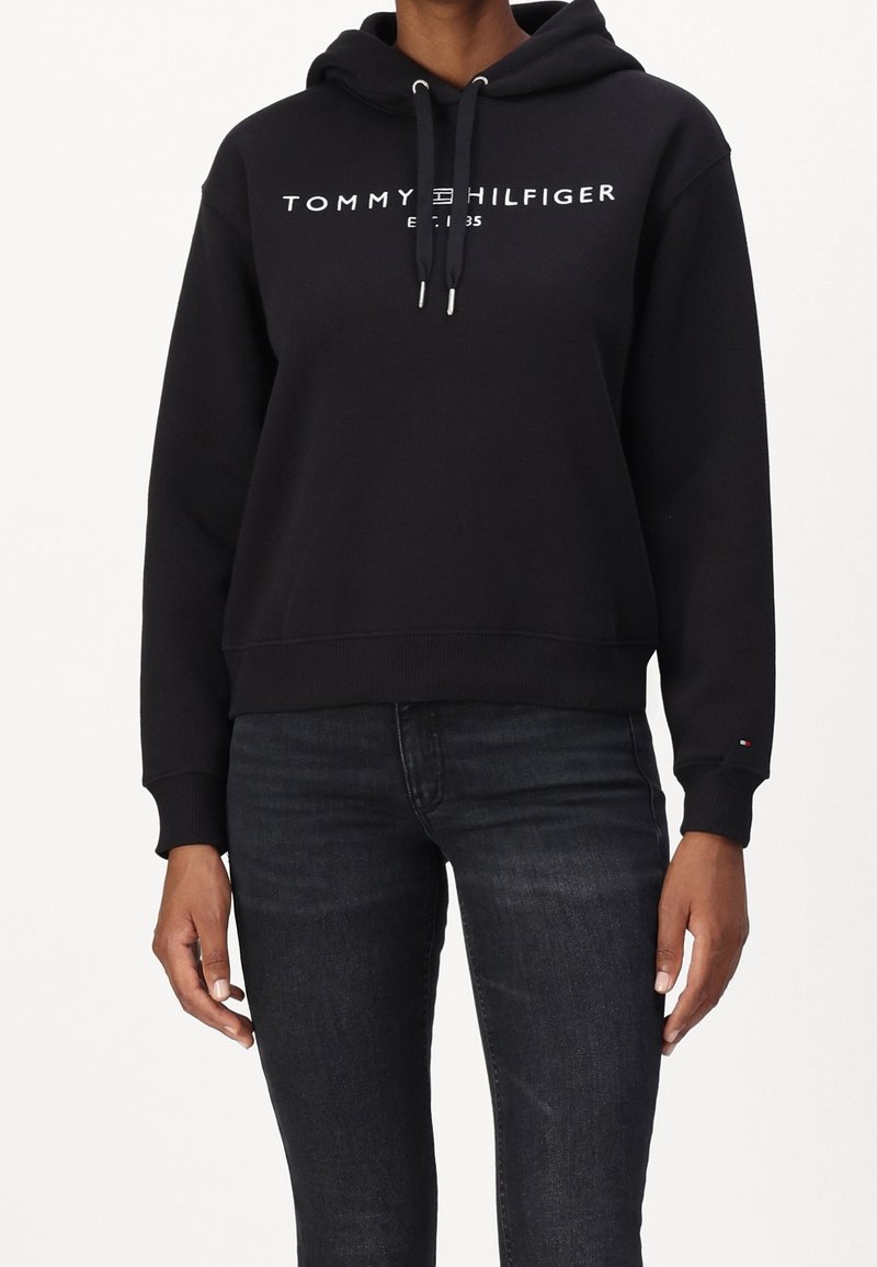 Schwarzer Kapuzenpullover mit weißem "TOMMY HILFIGER"-Text und Logo. Gerippte Bündchen und Saum. Getragen mit dunklen Jeans, die ein kleines Logo-Detail am Ärmel haben.