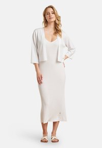 Witte cardigan met driekwartmouwen over een lichtbeige geribbelde gebreide jurk. De jurk is vloerlange met een lichte flair aan de onderkant.