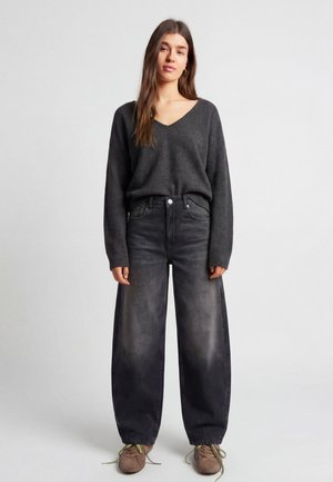 Nő áll, sötétszürke V-nyakú pulóvert, mosott fekete bő szárú farmert és bézs, zöld fűzőjű tornacipőt visel egyszínű háttér előtt.