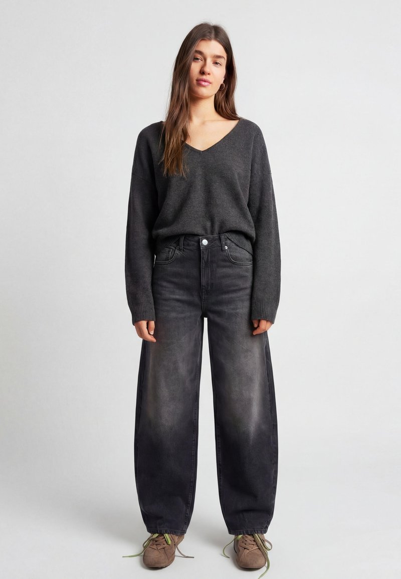 Femme debout, portant un pull à col en V gris foncé, un jean noir délavé à jambes larges et des baskets beiges avec des lacets verts, sur fond uni.