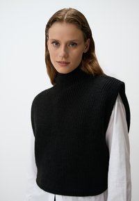 Zwarte gebreide mouwloze top met een hoge hals, voorzien van een geribbelde textuur en een cropped ontwerp, laag over een wit shirt.