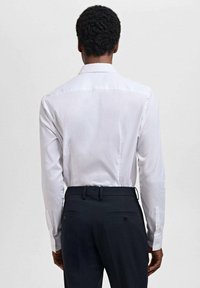 Camisa blanca de manga larga con una textura suave, mostrando un corte limpio y cuello clásico, combinada con pantalones oscuros que tienen bolsillos traseros.