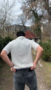 Ένα άτομο με λευκό πουκάμισο τραβάει ένα τηλέφωνο από την πίσω τσέπη του ενώ στέκεται σε έναν λιθόστρωτο δρόμο με θάμνους και κτίρια κοντά.