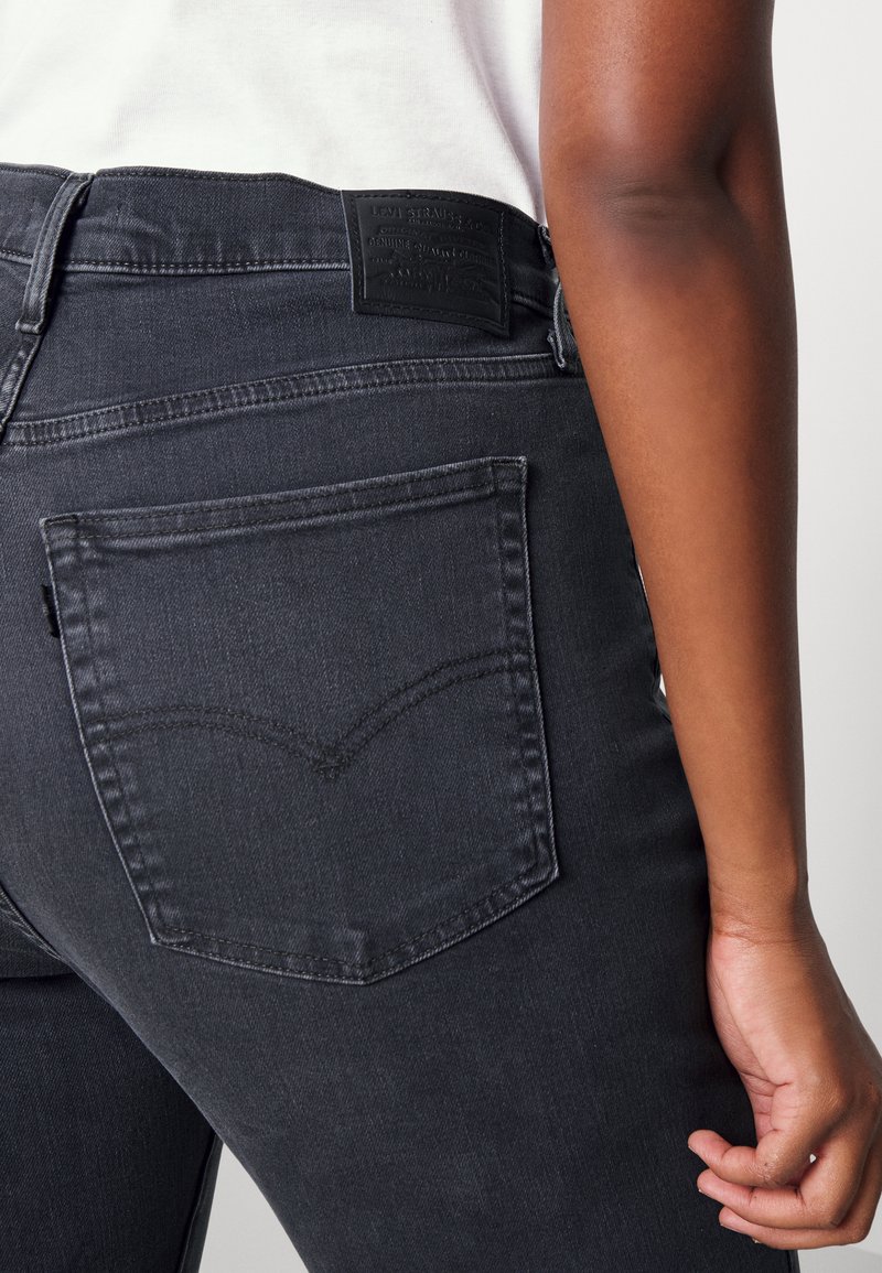 Dunkelgraue Denim-Jeans mit einer Gesäßtasche, die ein charakteristisches, geschwungenes Stichmuster aufweist, sowie einem schwarzen Leder-Logo-Patch am Bund.