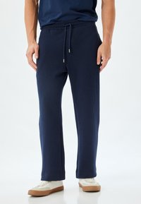 Pantalones de lounge de canalé azul marino con cinturilla elástica y cordón, con bolsillos laterales y un diseño de pierna recta. Se llevan zapatos blancos.