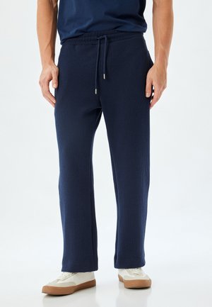 Pantalons de lounge côtelés bleu marine avec une taille élastique et un cordon de serrage, dotés de poches latérales et d'un design à jambes droites. Des chaussures blanches sont portées.