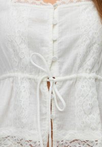 Blouse blanche avec des bordures en dentelle, dotée de boutons et d'une taille à cordon. Présente un tissu transparent et des détails en dentelle florale sur les côtés.