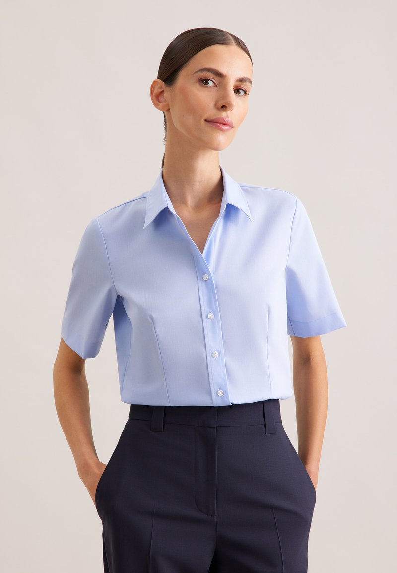 Camicia azzurro chiaro a maniche corte con bottoni e colletto appuntito, realizzata in tessuto liscio, abbinata a pantaloni scuri su misura.