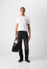 Fehér crew-neck póló kicsi piros és zöld logóval, fekete farmernadrág függőleges csíkokkal, fekete kézitáska, fekete cipő. Egyszerű háttér.