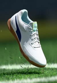 White athletic shoe with a gum sole, blue accent, and mesh details. Features a flat design and lace-up closure, hovering above grass.