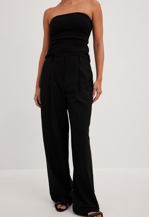 Femme portant un haut noir sans bretelles, un pantalon noir taille haute à jambes larges, et des sandales noires à bout ouvert sur un fond clair uni.